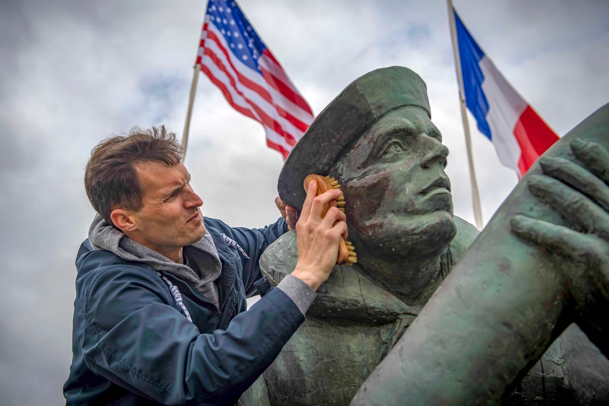 A sailor uses a brush to scrub the face of a statue outside, with a U.S. flag and a French flag in the background.