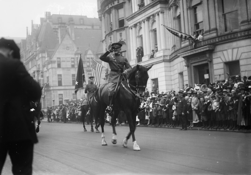 Army Gen. John J. Pershing salutes while parading on horseback on a city street filled with onlookers.