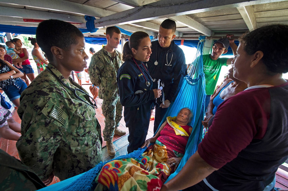 Several service members stand around a patient in a hammock.