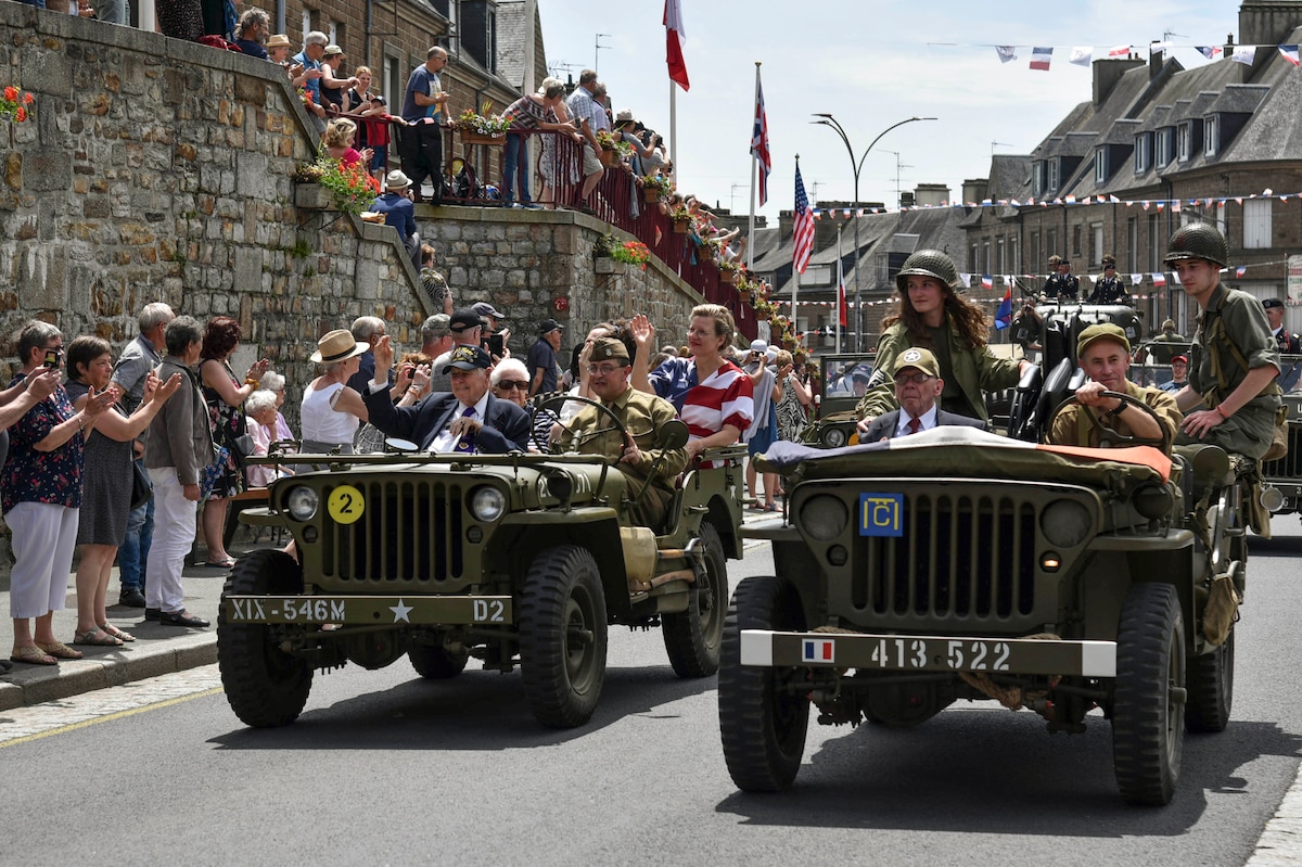 Veterans ride in jeeps down a village street where onlookers cheer.