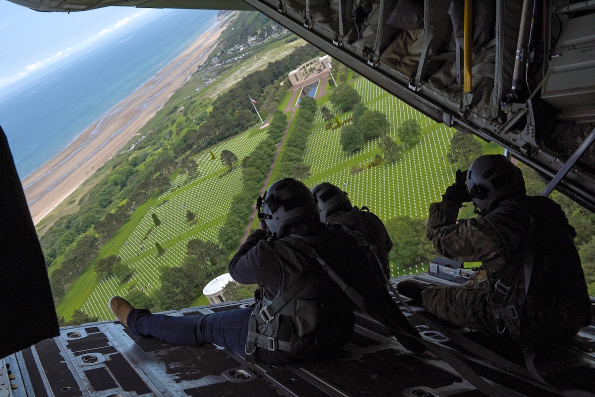 Three airmen, shown from behind, look out from an open aircraft over a cemetery by a shoreline.