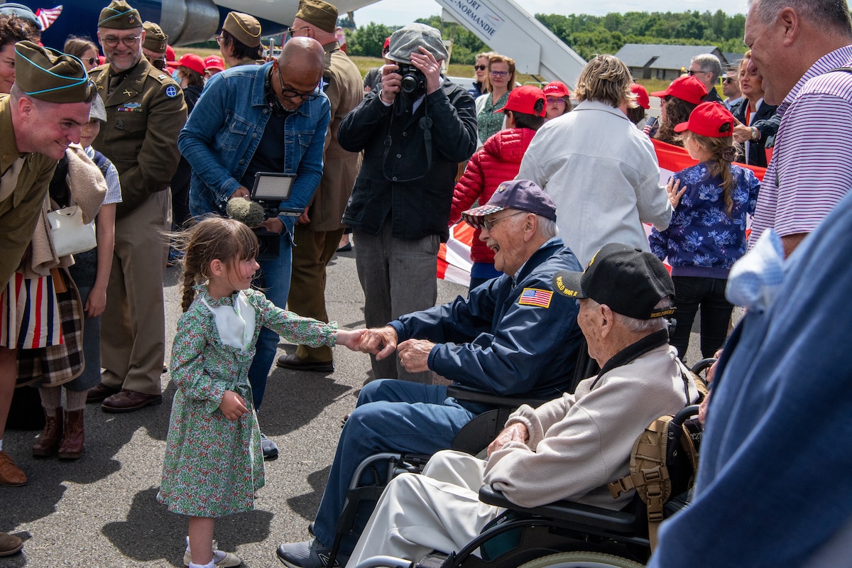 A child shakes hands with a veteran seated outside as other people look on and smile.