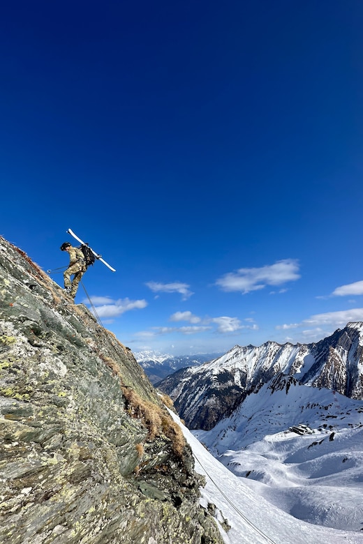 A soldier carrying skis attached to a backpack rappels on a rocky mountain face.