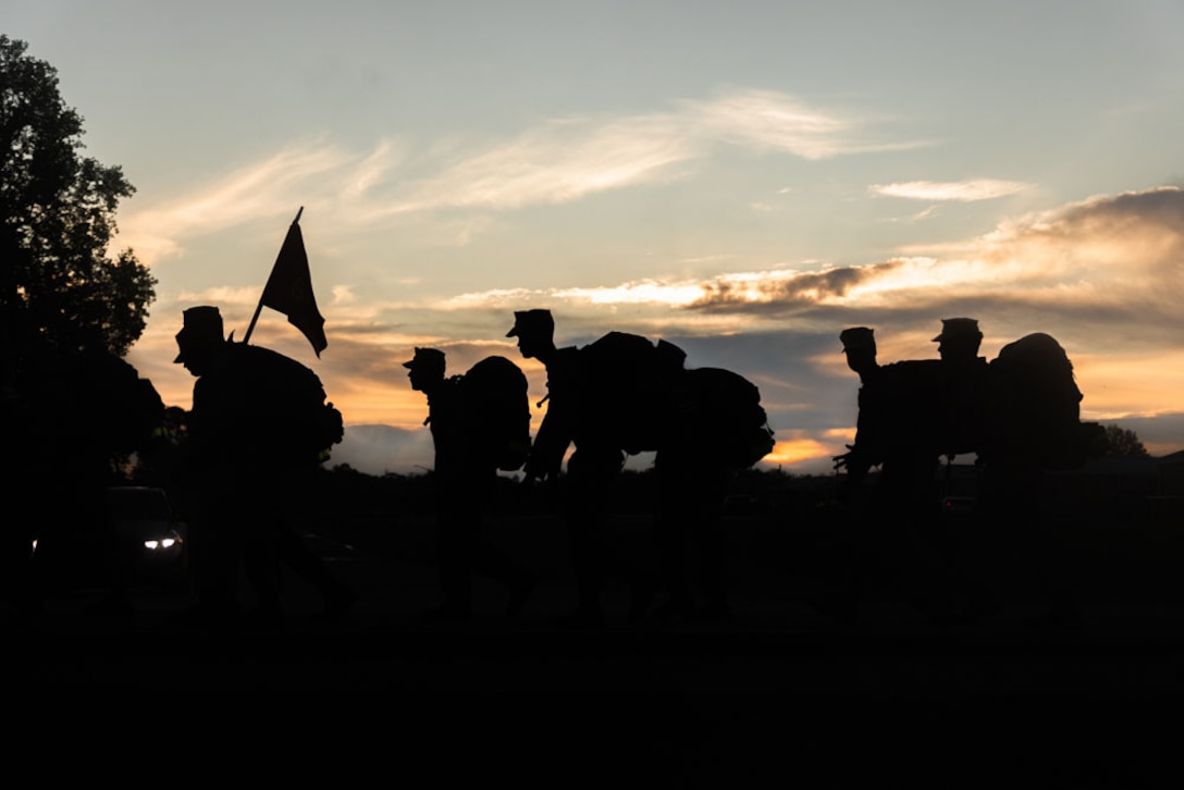 Marines walking and carrying backpacks are silhouetted against a twilight sky.