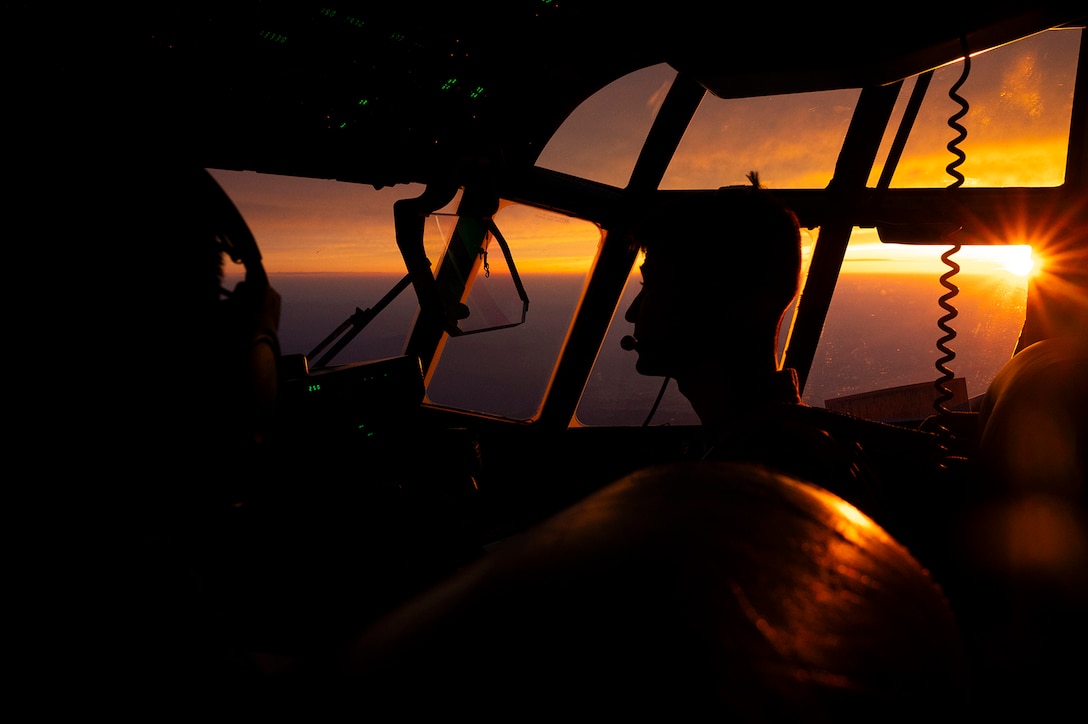 Silhouette view of a service member piloting a military aircraft with a low sun in the background.