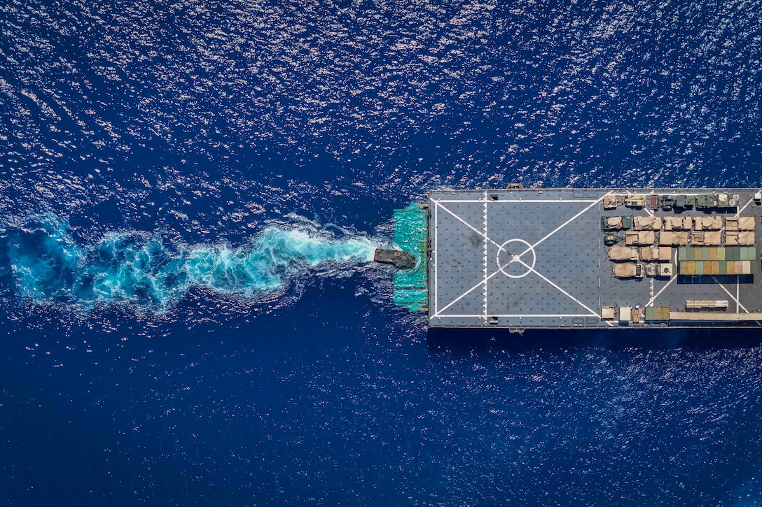 Aerial view of an amphibious combat vehicle approaching the landing deck of a Navy ship in open water.