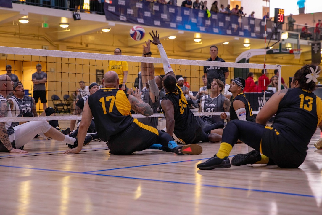 Athletes raise their hands to block a ball while sitting on opposite sides of a volleyball net as spectators watch from over a balcony.