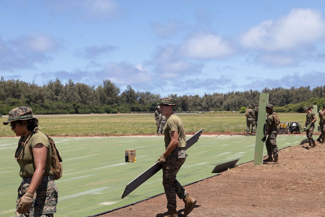 Marines prepare to lay more green panels on a helipad in a dirt area in a field.