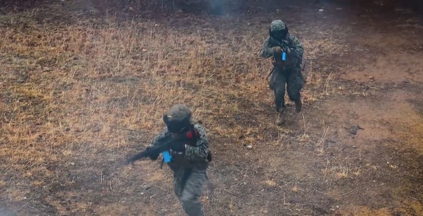Two Marines run through woods while carrying weapons.