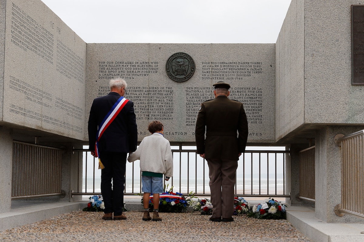A soldier, a civilian and a child, all shown from behind, stand at a concrete memorial on a beach, with wreaths at their feet.