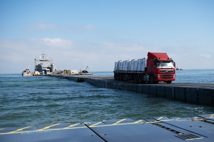 A truck carrying cargo drives on a pier away from a ship and toward land.