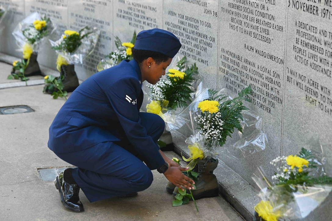 An airman places a yellow rose across the front of a pair of boots at a memorial. Other boots with yellow roses and bouquets are in the background.