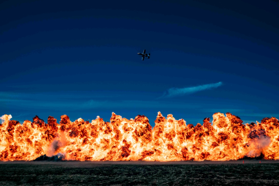 A military aircraft flies across a dark blue sky above a wall of flames.