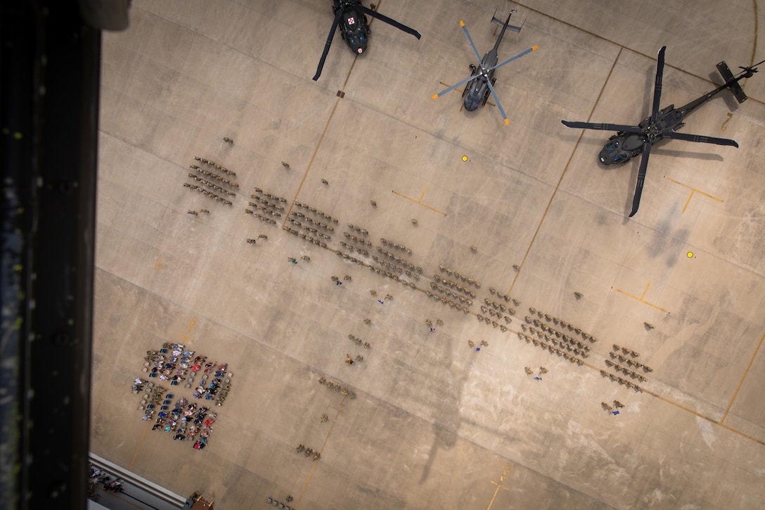 Dozens of soldiers stand in formation in front of three parked helicopters while a seated audience watches as seen from above.