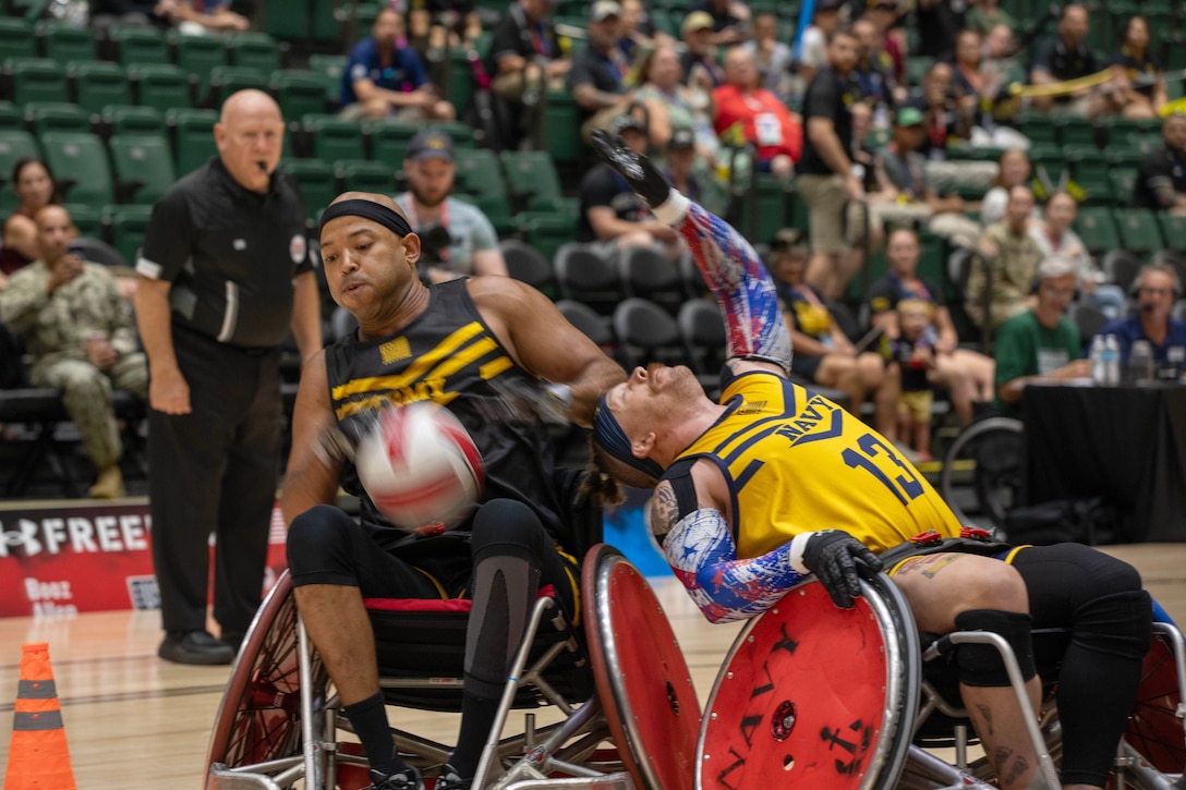 People in the stands and a referee on court watch an athlete in a wheelchair lean back into another aiming for a ball.