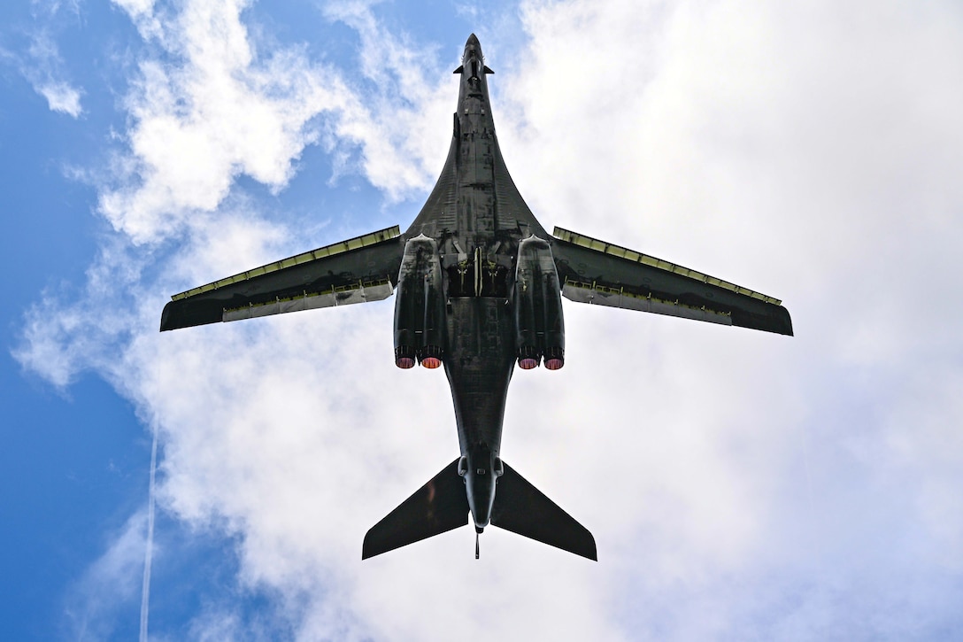 The bottom of a large military jet is seen from below against a partly cloudy sky.