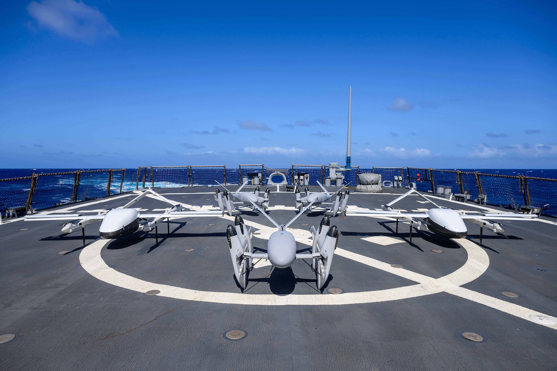 Five unmanned aerial systems sit on a ship’s flight deck while transiting a body of water.