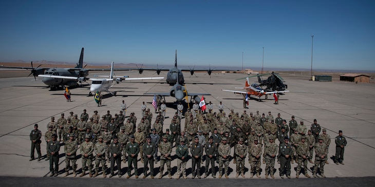 Representatives from the militaries of Peru, Colombia, Chile, Ecuador, Brazil, France and the United States pose for a group photo during the opening ceremony for Resolute Sentinel 2024 at Grupo 4 in La Joya, Peru, May 27, 2024, while military aircraft is shown behind and overhead.