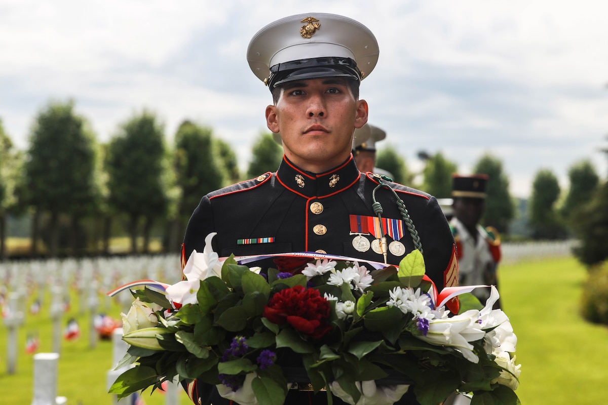 Medium close shot of a Marine in dress uniform holding a wreath and staring straight ahead with a backdrop of headstones in a cemetery.
